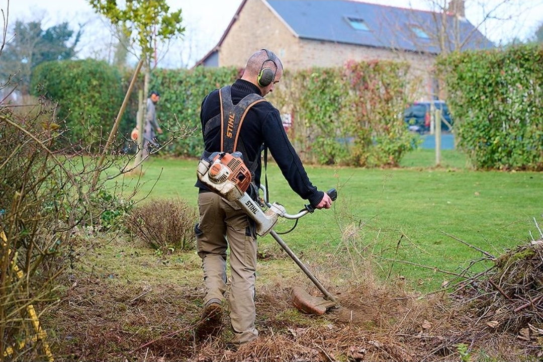 Jardinier utilisant une débroussailleuse thermique Stihl pour nettoyer des broussailles sèches et des herbes envahissantes en bordure de propriété
